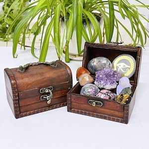 May include: Two small, wooden treasure chests, one closed and one open, displaying various colorful crystals and gemstones. The open chest reveals amethyst, agate, and other polished stones. The chests are brown with metal hardware, set against a backdrop of green foliage.