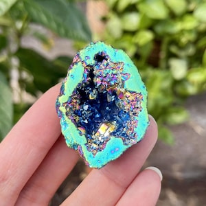 May include: A vibrant, iridescent geode with a teal and blue exterior. The center reveals a deep blue cavity filled with sparkling, multi-colored crystals. The geode is held in a hand, showcasing its natural, irregular shape.