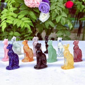 May include: A selection of small cat figurines in a range of colours, including pink, blue, orange, and green. The figurines are made from a translucent material and are arranged together. The background features green foliage and colourful flowers.