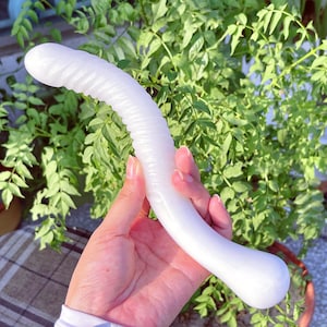 May include: A white, curved sex toy with a textured shaft and a smooth, rounded base. The toy is held against a backdrop of green foliage.