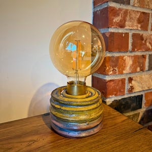 May include: A decorative table lamp with a large, round, amber-colored bulb. The base is a cylindrical shape with a textured, layered design in shades of yellow, blue, and purple. The lamp has a brass-colored neck.