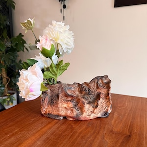 May include: A textured, rock-like ceramic vase in shades of brown and orange, featuring an irregular, rugged surface. The vase holds a small bouquet of artificial white and pale pink flowers with green leaves, arranged in a cluster on one side.