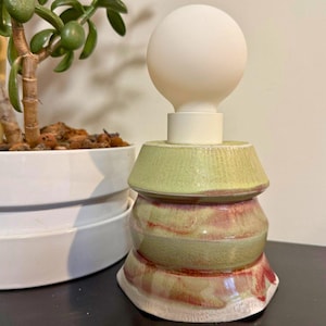 May include: A handmade ceramic lamp with a stacked design. The lamp base features a light green and red color scheme, topped with a white lightbulb. The lamp is a unique piece of home decor.