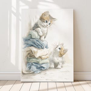 May include: A canvas print featuring two adorable kittens. One kitten sits atop a stack of folded pastel-colored linens, while the other looks up at it. The artwork has a soft, watercolor style, with a warm, inviting aesthetic.