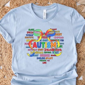 May include: A light blue t-shirt with the word "AUTISM" in colorful letters, surrounded by positive words like "unique" and "intelligent." The design includes a rainbow-colored ribbon. The shirt is made of soft material.