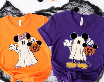 May include: An orange t-shirt featuring a ghost character with a pink polka dot bow and an orange pumpkin bucket, and a purple t-shirt featuring a ghost character with yellow shoes and an orange pumpkin bucket, both with short sleeves.