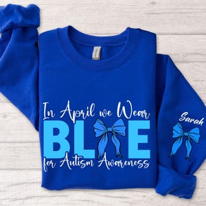 May include: Royal blue sweatshirt with the text "In April we Wear BLUE for Autism Awareness." The word "BLUE" is in a lighter shade of blue. A blue ribbon graphic is featured on the front and sleeve, with the name "Sarah" on the sleeve.