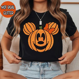 May include: A black short-sleeve t-shirt featuring a large orange pumpkin graphic shaped like a mouse head with ears and a bow. The graphic has a glitter-effect print and a jack-o'-lantern face. An orange circle in the corner says NOT REAL GLITTER.