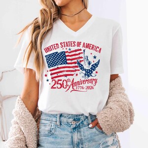 May include: White V-neck t-shirt with a distressed graphic of the American flag, the Statue of Liberty, and an eagle. The text reads "United States of America" and "250th Anniversary 1776-2026".