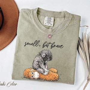 May include: Khaki-colored t-shirt with the text "small, but brave" and a graphic of a small monkey sitting on a stuffed animal. A silver necklace with a pendant is draped across the shirt.