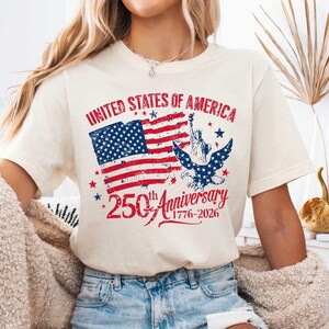 May include: Cream-coloured t-shirt with a distressed American flag design in red and blue. The shirt celebrates the 250th anniversary of the United States of America, with text reading "United States of America" and "250th Anniversary 1776-2026".