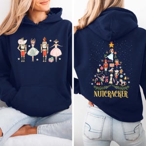 May include: A navy blue hooded sweatshirt featuring a front graphic of a mouse, ballerina, nutcracker, and dancer. The back displays a Christmas tree composed of various holiday icons and the text NUTCRACKER in yellow capital letters.