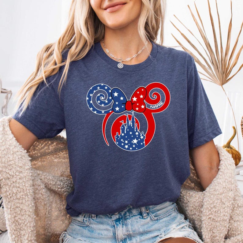 May include: Navy blue t-shirt featuring a patriotic design. The graphic includes a red and white bow, blue and white stars, and a castle silhouette. The word "NAME" is written on the right ear. A stylish and comfortable tee.