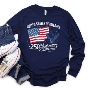 May include: Navy blue long-sleeved shirt featuring the text "UNITED STATES OF AMERICA" and "250th Anniversary 1776-2026". The graphic includes the American flag and an eagle. White trainers and blue jeans are visible.