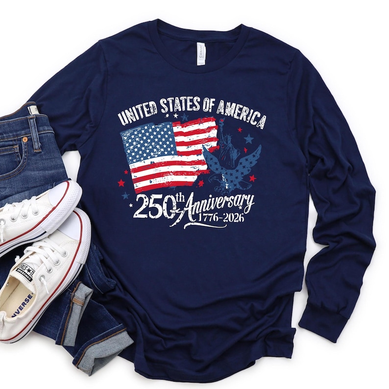 May include: Navy blue long-sleeved shirt featuring the text "UNITED STATES OF AMERICA" and "250th Anniversary 1776-2026". The graphic includes the American flag and an eagle. White trainers and blue jeans are visible.