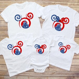 May include: White t-shirts and a onesie with a Mickey Mouse-inspired design. The design features a red and blue color scheme with a castle silhouette and star accents. The shirts are arranged on a wooden surface.