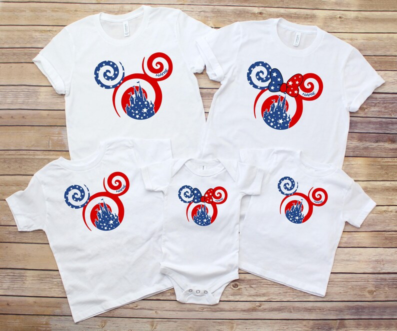 May include: White t-shirts and a onesie with a Mickey Mouse-inspired design. The design features a red and blue color scheme with a castle silhouette and star accents. The shirts are arranged on a wooden surface.