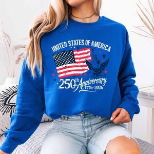 May include: A royal blue sweatshirt with the words "UNITED STATES OF AMERICA" above a graphic of the American flag and the Statue of Liberty. The text "250th Anniversary 1776-2026" is below the flag in white script.