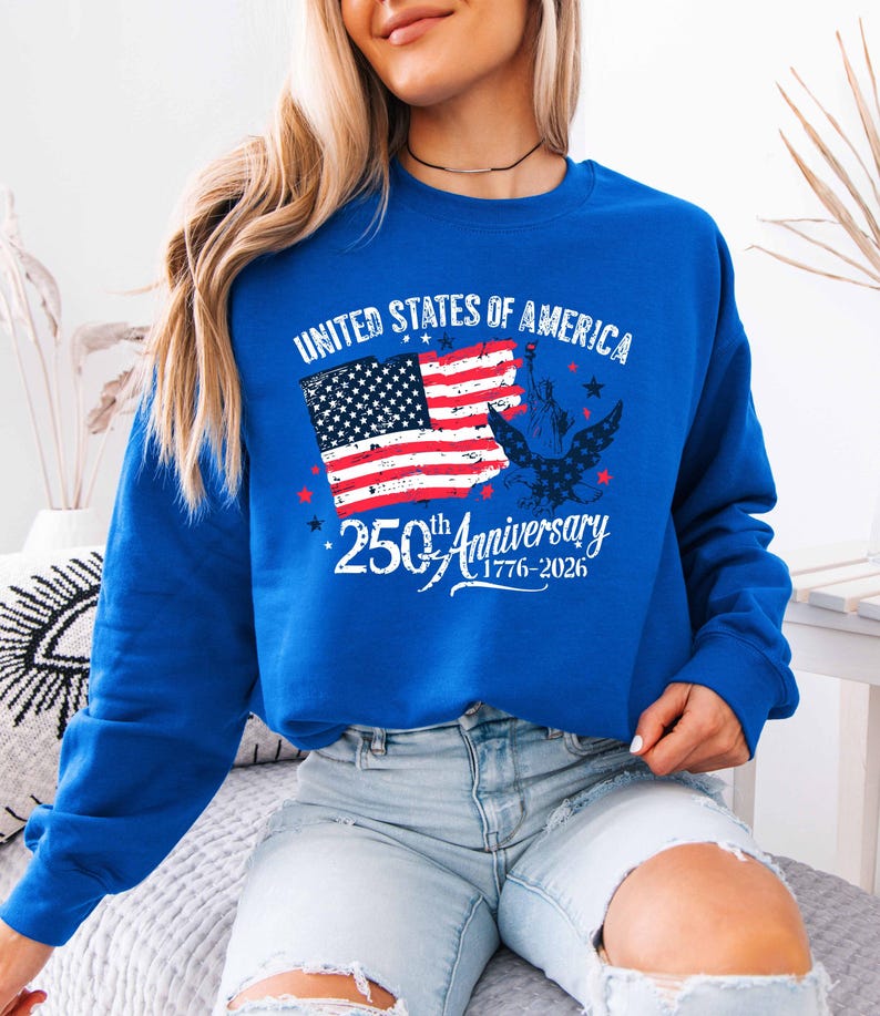 May include: A royal blue sweatshirt with the words "UNITED STATES OF AMERICA" above a graphic of the American flag and the Statue of Liberty. The text "250th Anniversary 1776-2026" is below the flag in white script.