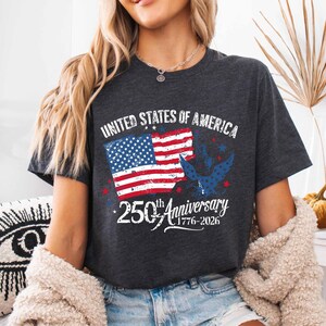 May include: Dark grey t-shirt with the text "UNITED STATES OF AMERICA" and a graphic of the American flag and a blue eagle. The shirt celebrates the 250th Anniversary, with the years 1776-2026.