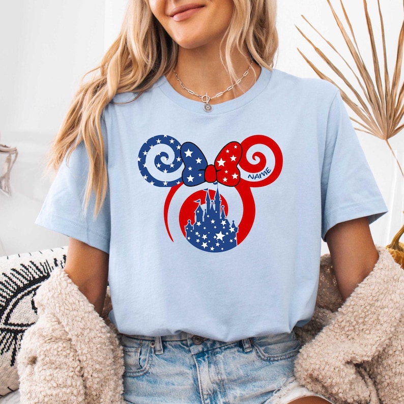 May include: Light blue t-shirt featuring a patriotic Mickey Mouse ear design. The design includes a red bow with the word "NAME", blue and white stars, and a castle silhouette. The shirt is a crew neck and has short sleeves.