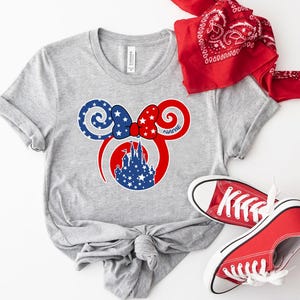 May include: A gray t-shirt featuring a patriotic design with a red, white, and blue Minnie Mouse ear graphic. The graphic includes a castle silhouette and stars. A red bandana and red sneakers complete the ensemble.