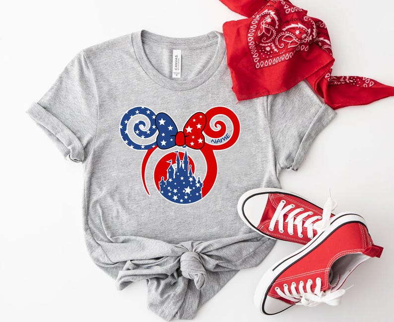 May include: A gray t-shirt featuring a patriotic design with a red, white, and blue Minnie Mouse ear graphic. The graphic includes a castle silhouette and stars. A red bandana and red sneakers complete the ensemble.