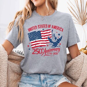 May include: A grey t-shirt featuring a patriotic design commemorating the 250th anniversary of the United States. The graphic includes the American flag, the Statue of Liberty, and the text "United States of America" and "250th Anniversary 1776-2026."