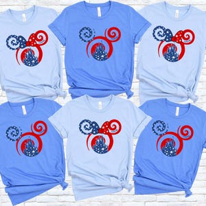 May include: Six light blue and blue t-shirts with a Mickey Mouse ear design. The design features a red bow, a castle, and stars. The shirts are displayed on a white brick wall.