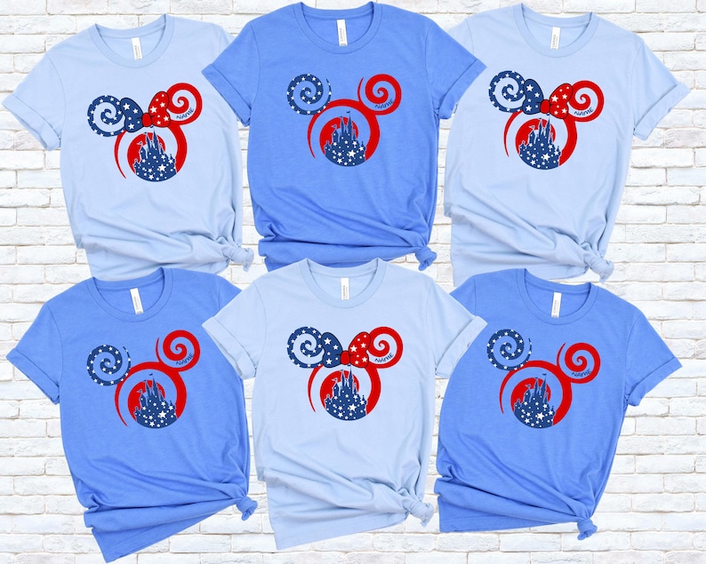May include: Six light blue and blue t-shirts with a Mickey Mouse ear design. The design features a red bow, a castle, and stars. The shirts are displayed on a white brick wall.