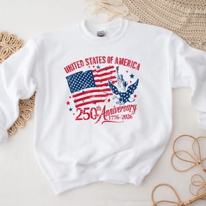 May include: White sweatshirt featuring a patriotic design commemorating the 250th anniversary of the United States. The design includes the American flag, an eagle, the Statue of Liberty, and the text "United States of America 250th Anniversary 1776-2026."