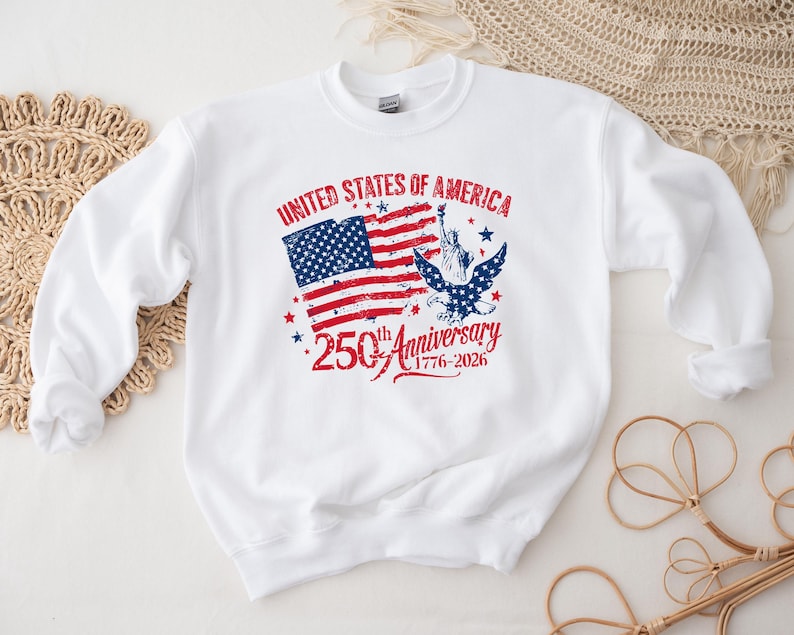 May include: White sweatshirt featuring a patriotic design commemorating the 250th anniversary of the United States. The design includes the American flag, an eagle, the Statue of Liberty, and the text "United States of America 250th Anniversary 1776-2026."