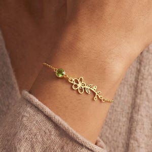 May include: A gold bracelet with a floral design and a round, light green gemstone. The bracelet has a delicate chain and is worn on the wrist. The floral design includes a flower and leaves.