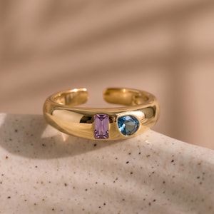 Op de afbeelding: Een goudkleurige ring met een open ontwerp, met een amethist en een blauwe edelsteen. De ring ligt op een wit oppervlak met een zachte, vage achtergrond. Het ontwerp van de ring is modern en elegant.