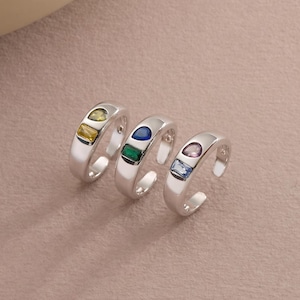 May include: Three silver-toned open rings, each featuring two small, distinctively shaped gemstones. The first ring has yellow and clear stones, the second has blue and green stones, and the third has purple and light blue stones.