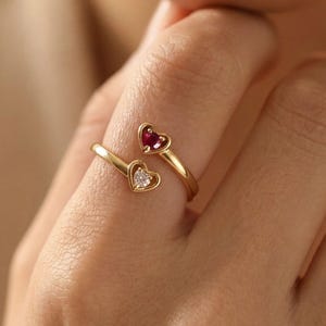 Può includere: Un anello dorato con due castoni a forma di cuore. Un cuore presenta una gemma rossa, l'altro una pietra trasparente e scintillante. L'anello è indossato su un dito, con i cuori rivolti in direzioni opposte.