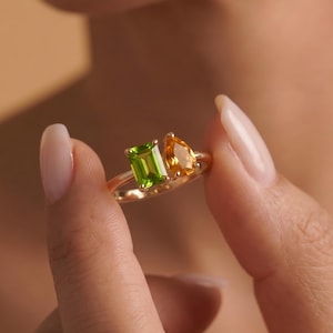 Op de afbeelding: Een gouden ring met een smaragdgeslepen groene peridoot en een peervormig geslepen gele citrien, naast elkaar geplaatst op een dunne gouden band. De tekst CustomMadeStoreGB is zichtbaar in de rechterbenedenhoek van de afbeelding.