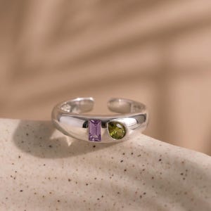 May include: A silver open-band ring featuring a rectangular purple gemstone and a teardrop-shaped green gemstone set side-by-side on the front. The band has a smooth, polished finish and a gap at the back, allowing for adjustable sizing.