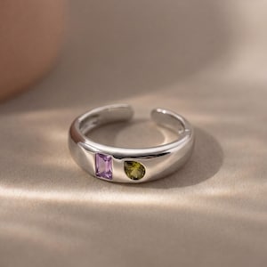 May include: A silver-toned open band ring featuring a smooth, polished finish. The ring is set with two small, distinct gemstones: one rectangular-cut light purple stone and one teardrop-shaped olive green stone, positioned side by side.