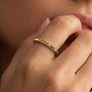 Può includere: Un anello a fascia color oro con una fila di cinque piccole pietre preziose rotonde e multicolori allineate. Le pietre presentano una sfumatura di colori che include rosa, giallo, azzurro, blu scuro e verde, incastonate nel metallo.