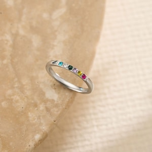 May include: A silver ring featuring a line of colourful gemstones. The gemstones are in shades of blue, green, pink, yellow, and red. The ring is displayed on a neutral-toned surface.