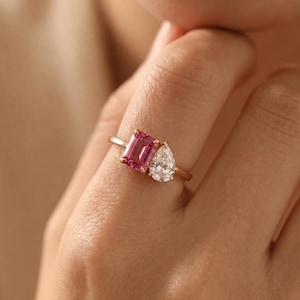 Peut inclure: Une bague en or avec deux pierres serties côte à côte sur un anneau fin. Une pierre est une gemme rose de taille émeraude rectangulaire, et l'autre est une gemme blanche transparente en forme de poire. Le texte CustomMadeStoreGB est visible.