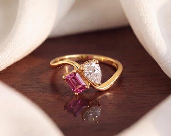 May include: A gold ring featuring a bypass band design with two prominent gemstones. One stone is a rectangular emerald-cut pink gemstone, and the other is a pear-shaped clear white gemstone. The ring has a polished finish and a reflective surface.