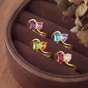 Könnte beinhalten: Vier Goldringe, jeder mit einem Design aus zwei Edelsteinen, bestehend aus einem Smaragdschliff-Edelstein und einem tropfenförmigen Edelstein. Die Ringe zeigen verschiedene Farbkombinationen, darunter violette, blaue, grüne und rote Steine.