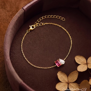 Puede incluir: Una delicada pulsera de cadena dorada que presenta dos piedras prominentes: una gema de color rosa vibrante con corte rectangular y una gema blanca transparente con forma de pera. Incluye una cadena fina de eslabones con cierre de mosquetón y extensión.