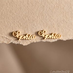 Op de afbeelding: Een paar goudkleurige metalen oorknopjes, elk met de naam Sophia geschreven in een cursief lettertype. De letters zijn verbonden in één stuk, wat zorgt voor een gepolijste, metalen afwerking die het licht op het oppervlak weerkaatst.