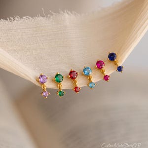 Pode incluir: Um conjunto de seis brincos pendentes dourados, cada um com duas pedras preciosas de corte redondo em cores vibrantes, incluindo roxo, verde, vermelho, azul claro, magenta e azul escuro, dispostos em fila sobre um tecido claro texturizado.