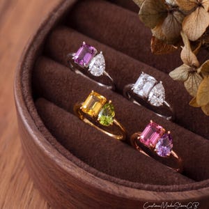 May include: Four rings arranged in a brown velvet display case. Each ring features a dual-stone design with one emerald-cut gemstone and one pear-cut gemstone. The rings include combinations of pink, clear, yellow, green, and purple stones in metal bands.