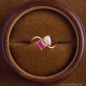 Könnte beinhalten: Ein goldener Ring mit einem Bypass-Design, besetzt mit einem rechteckigen rosa Edelstein und einem birnenförmigen klaren Edelstein. Der Ring liegt in einer runden Holzschatulle mit braunem Samtinnenfutter. Der Text CustomMadeStoreGB ist unten zu sehen.