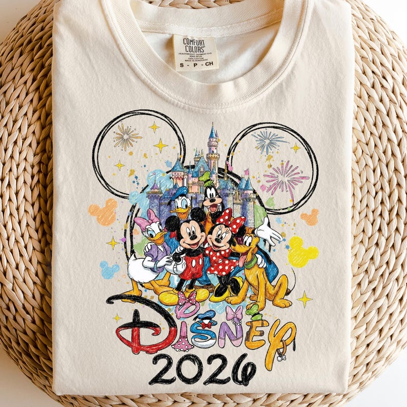 K&ouml;nnte beinhalten: Cremefarbenes T-Shirt mit einer farbenfrohen Grafik von Disney-Figuren, darunter Micky und Minnie Maus, Donald Duck und Goofy. Das Design umfasst ein Schloss, Feuerwerk und den Text "Disney 2026."
